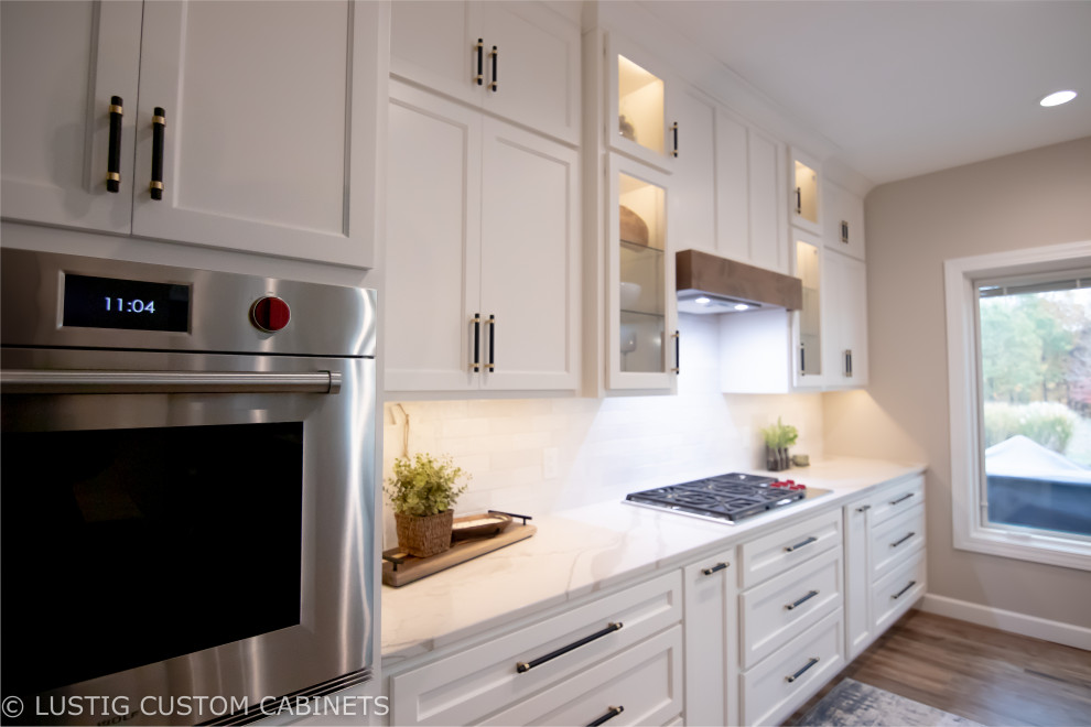 Large transitional l-shaped laminate floor and brown floor open concept kitchen photo with an undermount sink, flat-panel cabinets, white cabinets, quartz countertops, white backsplash, ceramic backsplash, stainless steel appliances, an island and white countertops