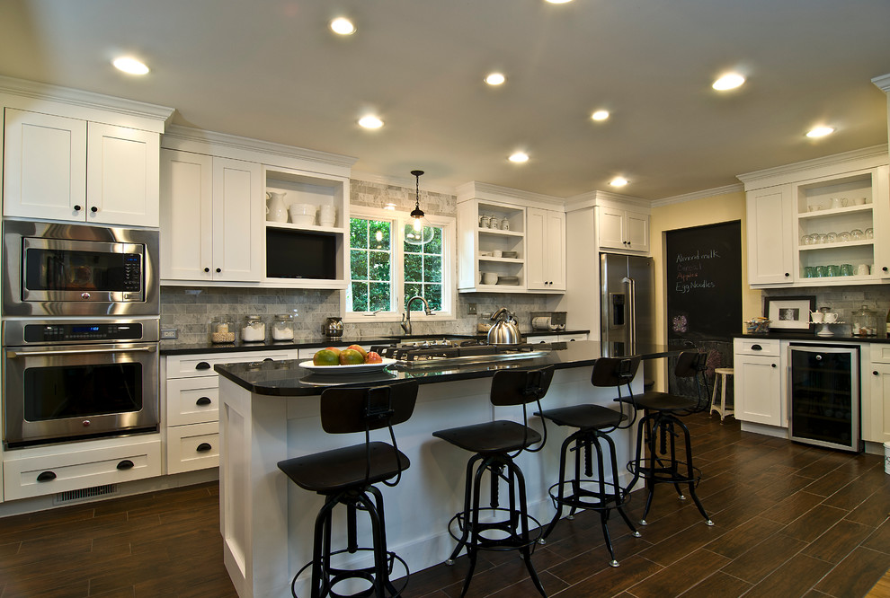 This is an example of a large transitional l-shaped kitchen in Raleigh with a farmhouse sink, shaker cabinets, white cabinets, granite benchtops, grey splashback, stone tile splashback, stainless steel appliances, dark hardwood floors, with island and brown floor.