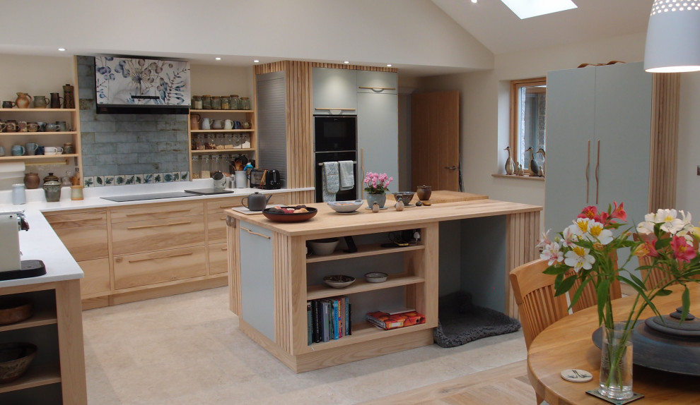 This is an example of a large contemporary l-shaped open plan kitchen in Cornwall with a submerged sink, flat-panel cabinets, light wood cabinets, engineered stone countertops, blue splashback, black appliances, limestone flooring, an island, beige floors, white worktops, exposed beams and feature lighting.