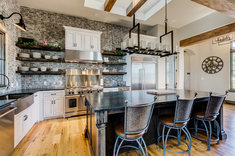 This is an example of a mid-sized industrial l-shaped open plan kitchen in Dallas with a farmhouse sink, recessed-panel cabinets, white cabinets, granite benchtops, grey splashback, subway tile splashback, stainless steel appliances, light hardwood floors, with island, brown floor and black benchtop.