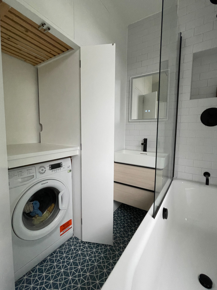 This is an example of a small contemporary family bathroom in London with flat-panel cabinets, light wood cabinets, a built-in bath, a shower/bath combination, a two-piece toilet, white tiles, porcelain tiles, white walls, porcelain flooring, a wall-mounted sink, blue floors, a hinged door, a laundry area, a single sink and a floating vanity unit.