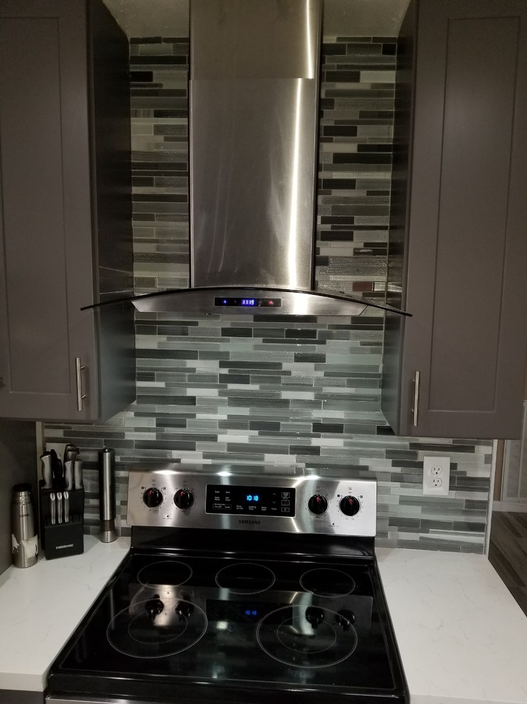 Mid-sized contemporary l-shaped eat-in kitchen in Houston with a farmhouse sink, shaker cabinets, grey cabinets, quartzite benchtops, grey splashback, glass sheet splashback, stainless steel appliances, porcelain floors, with island, grey floor and white benchtop.