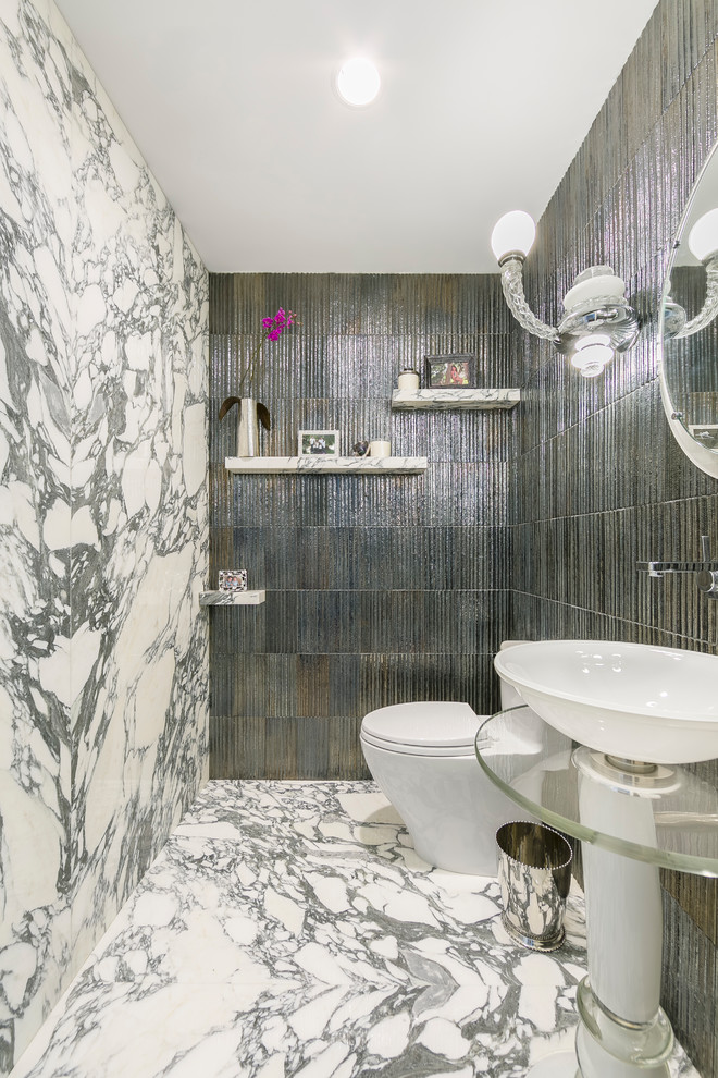 This is an example of an eclectic powder room in New York with glass-front cabinets, a one-piece toilet, multi-coloured tile, stone slab, multi-coloured walls, marble floors, a pedestal sink, glass benchtops and multi-coloured floor.