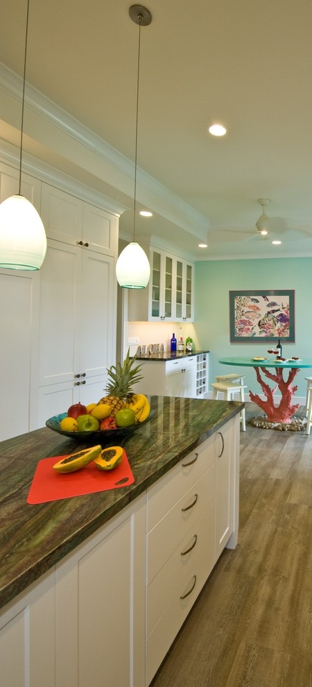 Photo of a tropical kitchen in Hawaii with a farmhouse sink, shaker cabinets, white cabinets, granite benchtops, green splashback, glass tile splashback, panelled appliances, vinyl floors and with island.