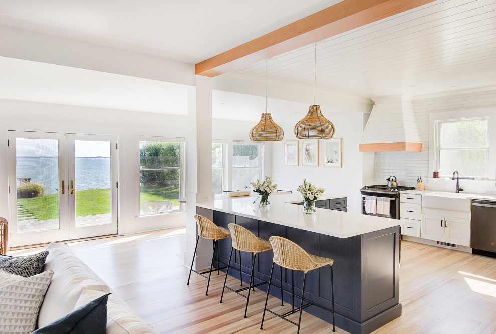 Photo of a mid-sized beach style open plan kitchen in New York with a farmhouse sink, recessed-panel cabinets, blue cabinets, quartz benchtops, white splashback, ceramic splashback, stainless steel appliances, light hardwood floors, with island, white benchtop and timber.