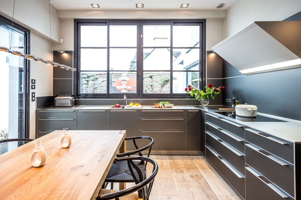 This is an example of a mid-sized contemporary l-shaped eat-in kitchen in Brest with flat-panel cabinets, grey cabinets, no island, a single-bowl sink, stainless steel benchtops, black splashback, panelled appliances, medium hardwood floors, brown floor and grey benchtop.