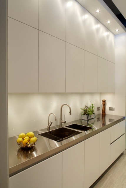 This is an example of a modern single-wall separate kitchen in London with an integrated sink, flat-panel cabinets, white cabinets, stainless steel benchtops, white splashback and stainless steel appliances.