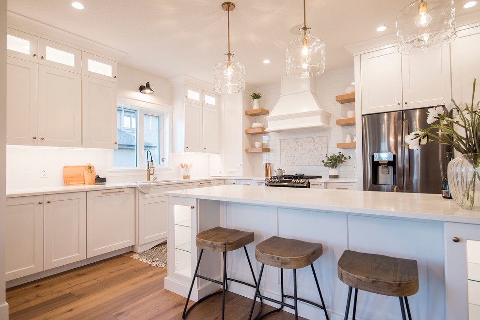 Mid-sized transitional l-shaped kitchen pantry in Edmonton with a drop-in sink, shaker cabinets, white cabinets, quartzite benchtops, white splashback, subway tile splashback, stainless steel appliances, medium hardwood floors, with island, brown floor and white benchtop.