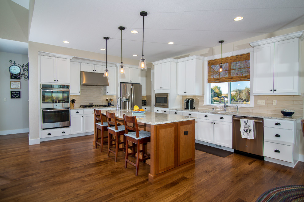 This is an example of a large arts and crafts l-shaped open plan kitchen in Denver with an undermount sink, shaker cabinets, white cabinets, granite benchtops, beige splashback, glass tile splashback, stainless steel appliances, medium hardwood floors, with island and brown floor.