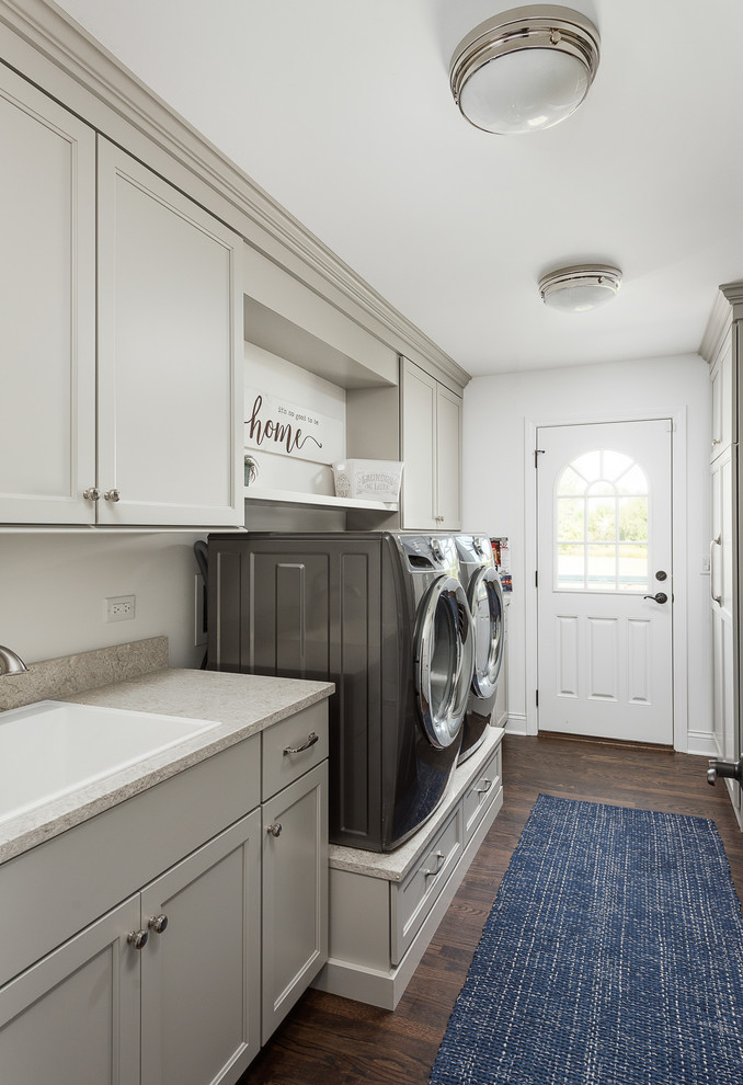 Photo of a large transitional galley utility room in Chicago with a drop-in sink, flat-panel cabinets, white cabinets, quartz benchtops, grey walls, dark hardwood floors, a side-by-side washer and dryer, brown floor and grey benchtop.