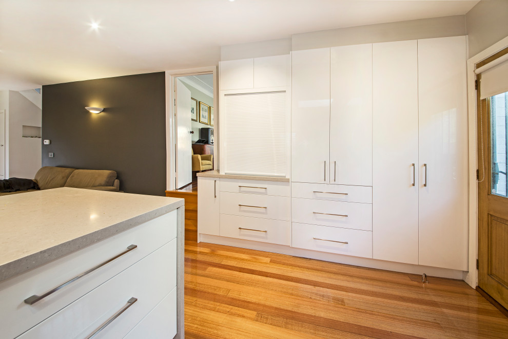 Inspiration for a large modern l-shaped kitchen pantry in Melbourne with an undermount sink, flat-panel cabinets, white cabinets, quartz benchtops, metallic splashback, glass sheet splashback, stainless steel appliances, light hardwood floors, with island and beige benchtop.
