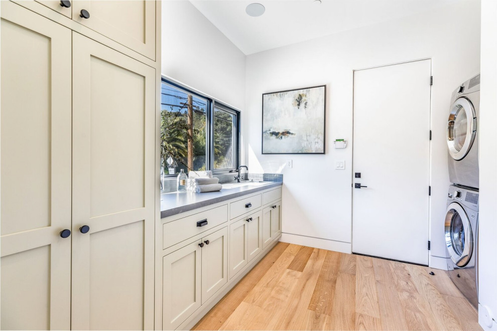 Photo of a large contemporary galley dedicated laundry room in Los Angeles with an undermount sink, shaker cabinets, white cabinets, quartz benchtops, grey splashback, mosaic tile splashback, white walls, light hardwood floors, a stacked washer and dryer, beige floor and grey benchtop.