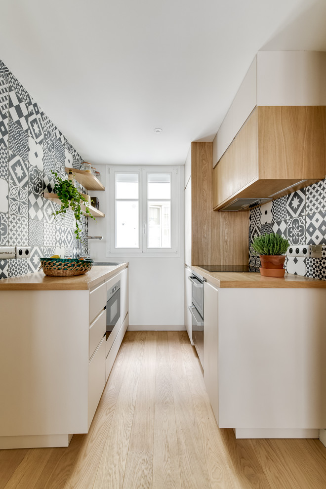 This is an example of a scandinavian galley kitchen in Paris with flat-panel cabinets, white cabinets, wood benchtops, multi-coloured splashback, cement tile splashback, black appliances, light hardwood floors, no island and beige floor.