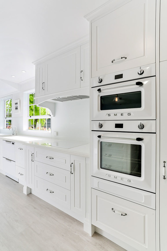 Inspiration for a mid-sized country u-shaped eat-in kitchen in Other with an undermount sink, recessed-panel cabinets, white cabinets, solid surface benchtops, white splashback, glass sheet splashback, white appliances, vinyl floors, no island, brown floor and white benchtop.