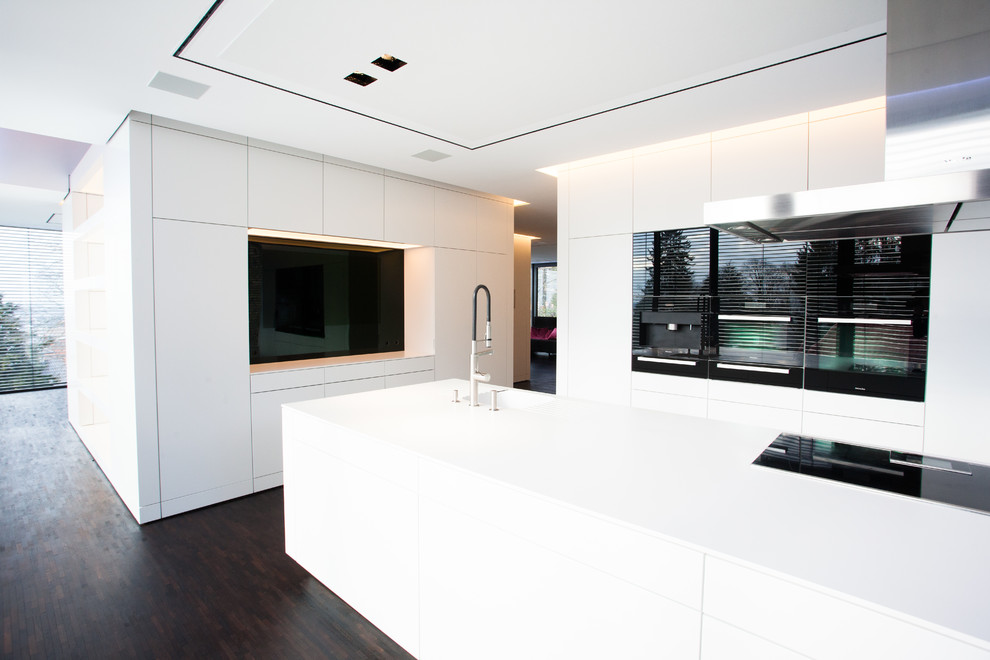 This is an example of an expansive modern open plan kitchen in Frankfurt with an integrated sink, flat-panel cabinets, white cabinets, black splashback, glass sheet splashback, black appliances, dark hardwood floors and with island.