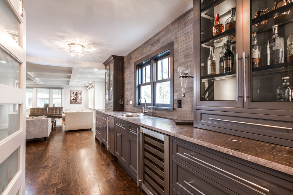 Photo of a mid-sized contemporary single-wall wet bar in Indianapolis with a drop-in sink, beaded inset cabinets, marble benchtops, porcelain splashback, medium hardwood floors, brown floor, brown cabinets and brown splashback.
