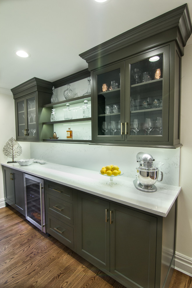 This is an example of a small transitional l-shaped wet bar in New York with no sink, flat-panel cabinets, green cabinets, quartz benchtops, white splashback, stone slab splashback, dark hardwood floors, brown floor and white benchtop.