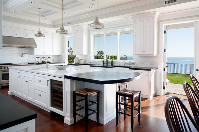 This is an example of a large beach style u-shaped eat-in kitchen in Los Angeles with with island, a farmhouse sink, raised-panel cabinets, white cabinets, recycled glass benchtops, white splashback, stone tile splashback, stainless steel appliances and medium hardwood floors.