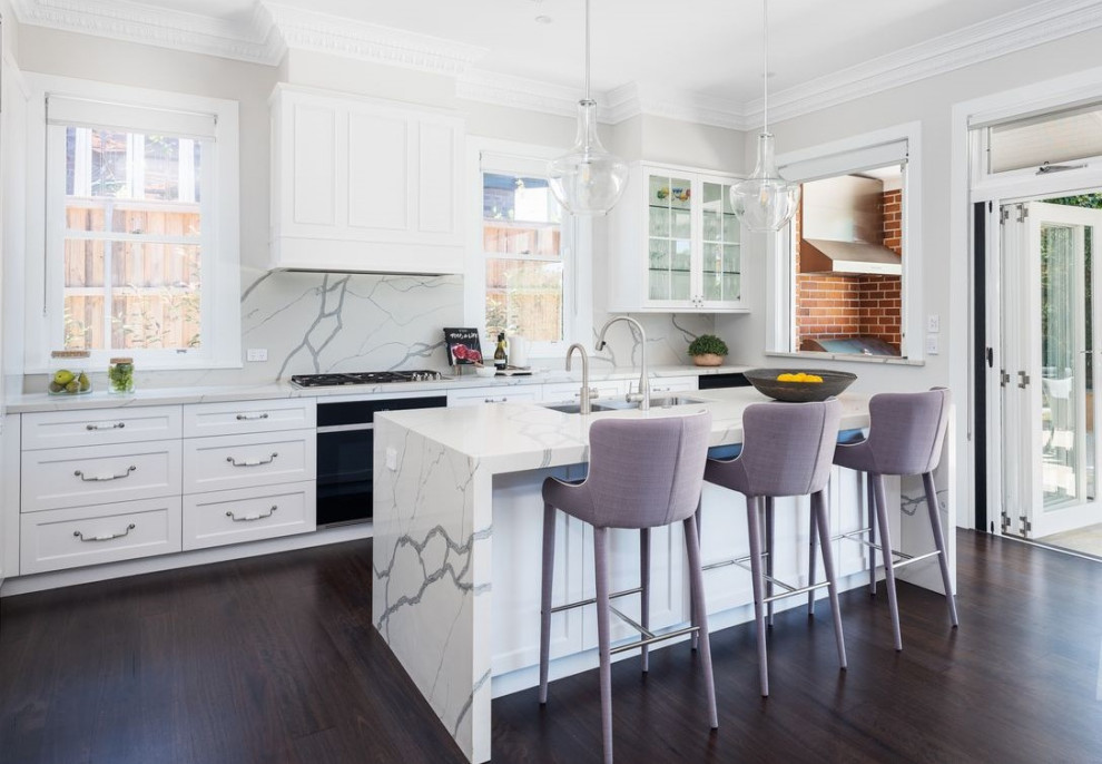 Design ideas for a mid-sized traditional l-shaped open plan kitchen in Sydney with an undermount sink, shaker cabinets, white cabinets, marble benchtops, white splashback, marble splashback, black appliances, dark hardwood floors, with island, brown floor and white benchtop.