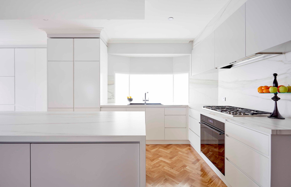 Modern l-shaped kitchen in Melbourne with a drop-in sink, flat-panel cabinets, white cabinets, white splashback, stone slab splashback, black appliances, medium hardwood floors, with island, brown floor and white benchtop.