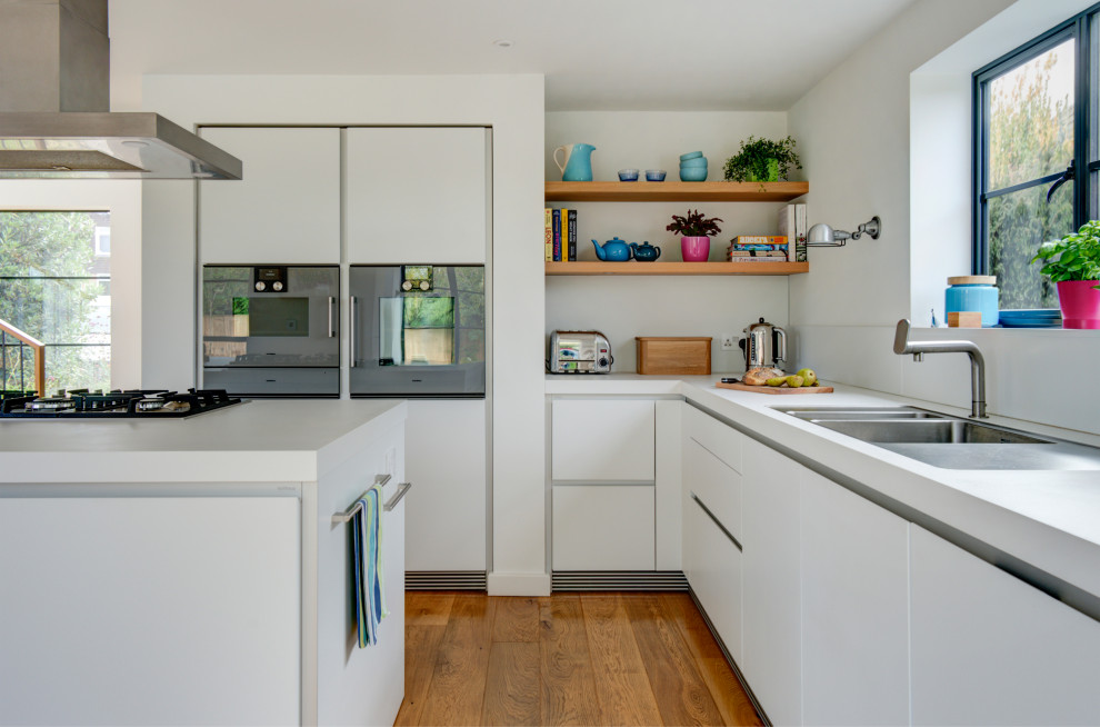 Photo of a contemporary l-shaped kitchen/diner in Gloucestershire with an integrated sink, flat-panel cabinets, white cabinets, laminate countertops, white splashback, glass sheet splashback, black appliances, medium hardwood flooring, an island and white worktops.