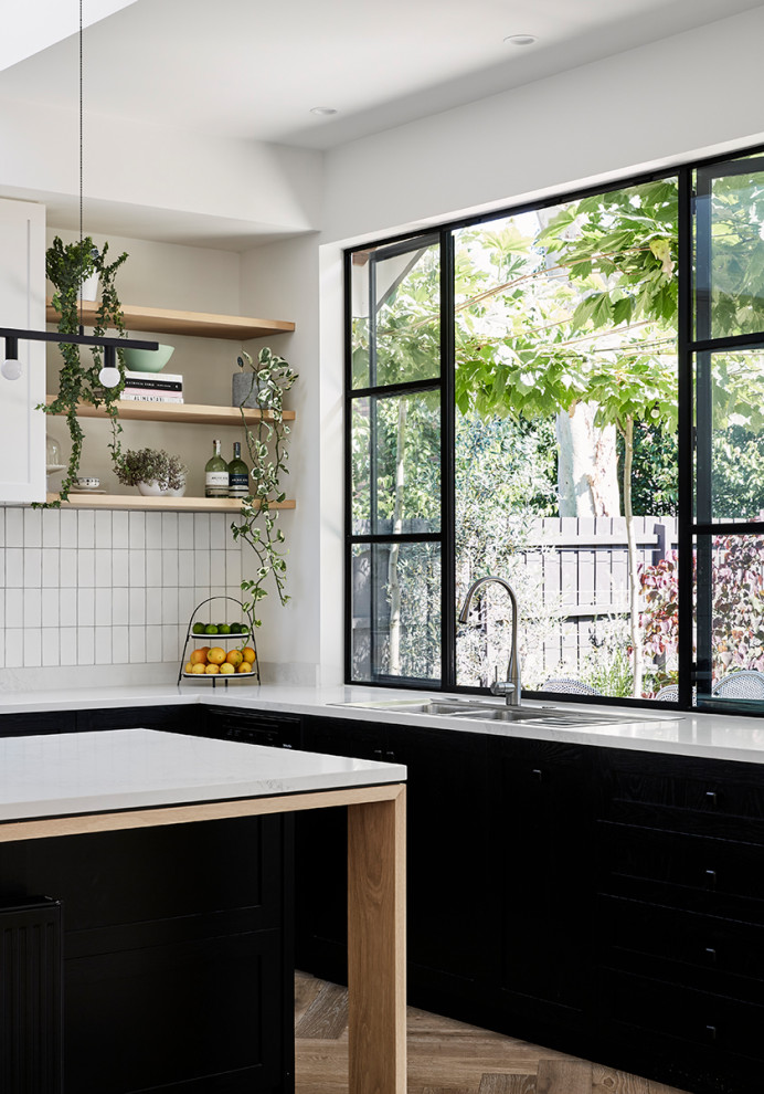 Photo of a mid-sized traditional u-shaped open plan kitchen in Melbourne with shaker cabinets, black cabinets, quartz benchtops, white splashback, ceramic splashback, black appliances, light hardwood floors, with island, brown floor and white benchtop.