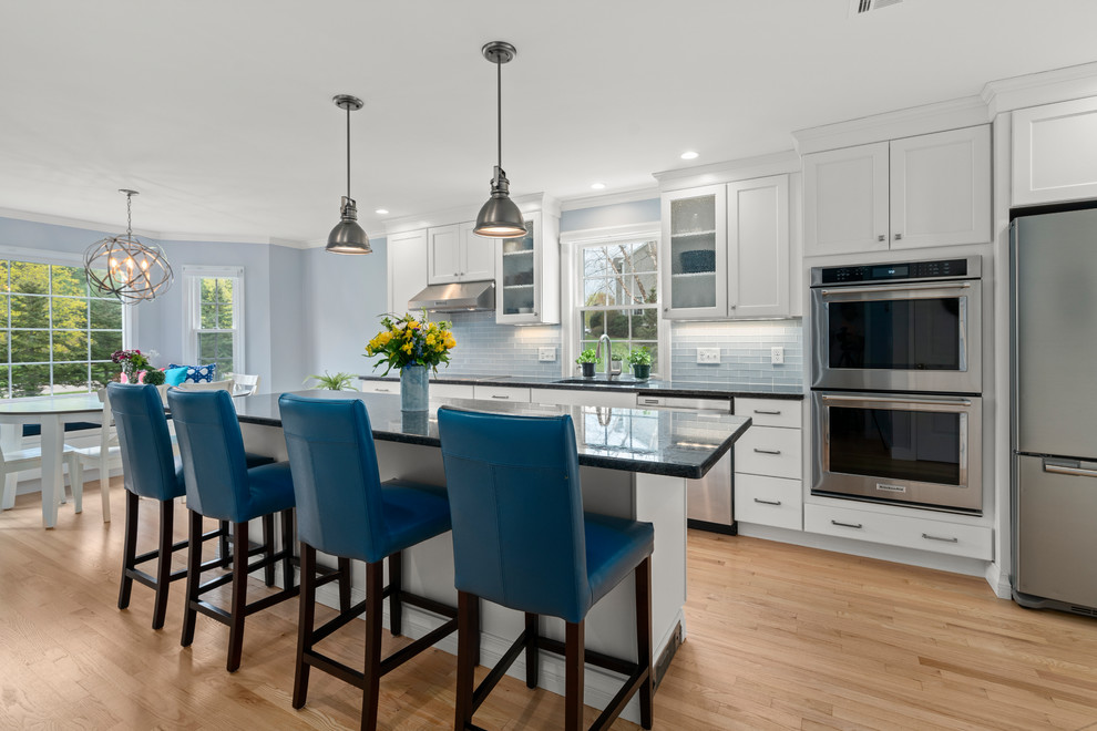 This is an example of a transitional galley kitchen in Boston with an undermount sink, shaker cabinets, white cabinets, blue splashback, stainless steel appliances, light hardwood floors, with island, beige floor and black benchtop.