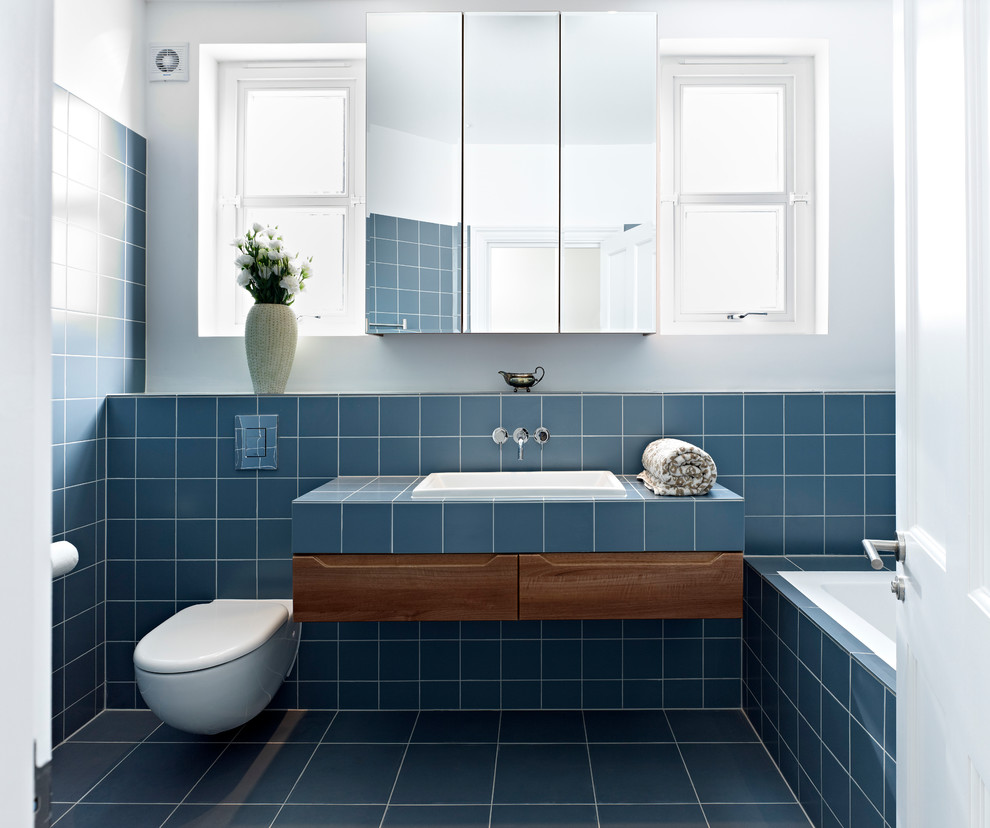 This is an example of a mid-sized contemporary bathroom in London with medium wood cabinets, tile benchtops, a drop-in tub, a drop-in sink, a wall-mount toilet, blue tile, white walls and flat-panel cabinets.