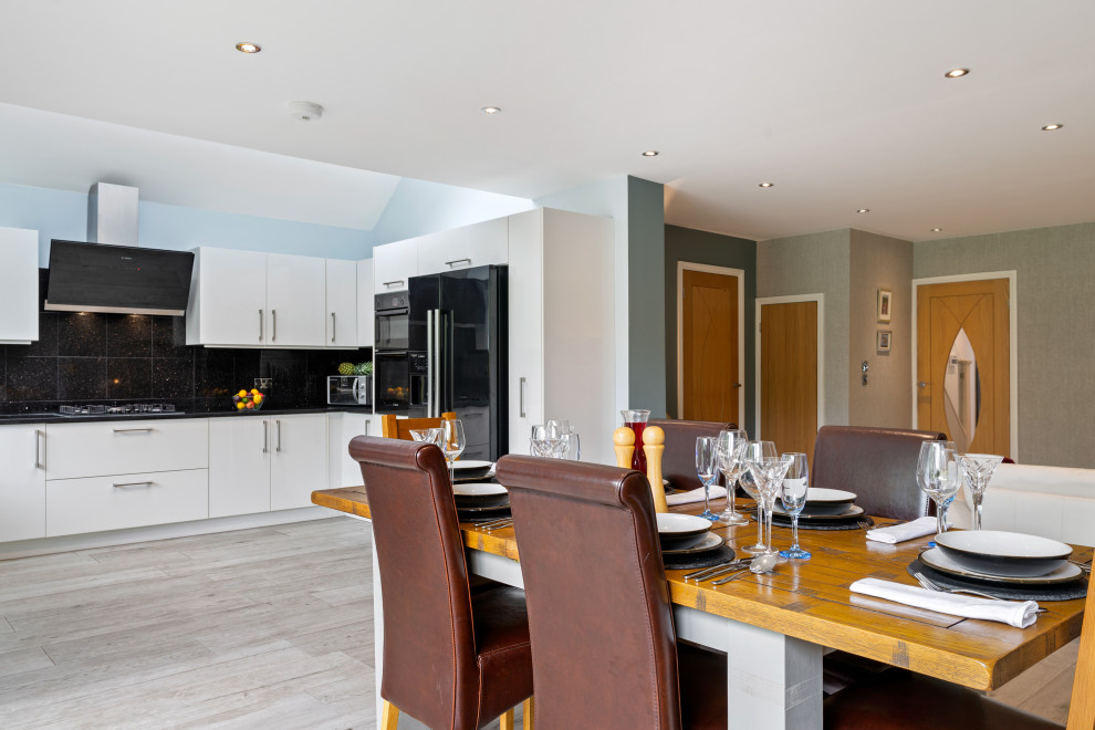 This is an example of a mid-sized contemporary l-shaped open plan kitchen in Surrey with a drop-in sink, flat-panel cabinets, white cabinets, quartzite benchtops, black splashback, engineered quartz splashback, black appliances, porcelain floors, grey floor and black benchtop.