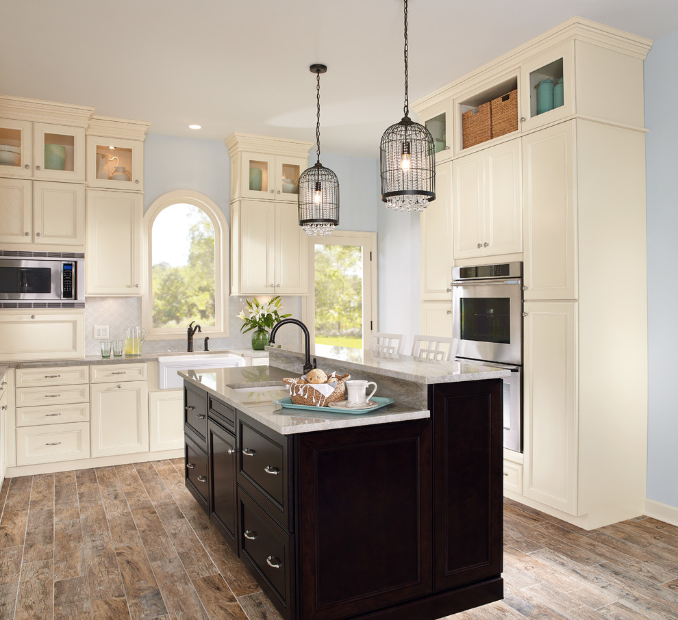 Mid-sized transitional u-shaped medium tone wood floor and brown floor enclosed kitchen photo with a farmhouse sink, recessed-panel cabinets, white cabinets, quartzite countertops, gray backsplash, porcelain backsplash, stainless steel appliances and an island