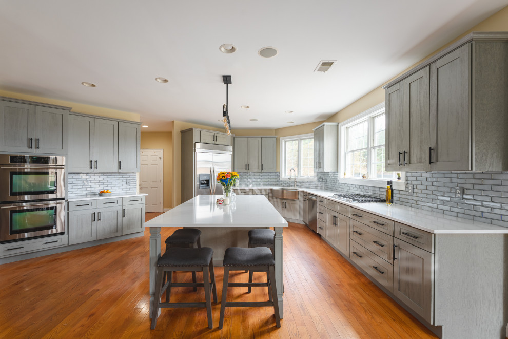 Inspiration for a large transitional u-shaped medium tone wood floor and brown floor eat-in kitchen remodel with a farmhouse sink, shaker cabinets, gray cabinets, quartz countertops, gray backsplash, glass tile backsplash, stainless steel appliances, an island and white countertops