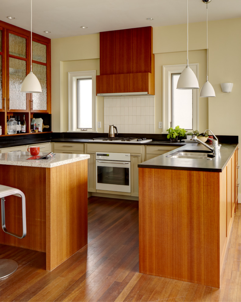 This is an example of a transitional u-shaped kitchen in Seattle with an undermount sink, shaker cabinets, beige cabinets, white splashback, white appliances, medium hardwood floors, with island, brown floor and black benchtop.