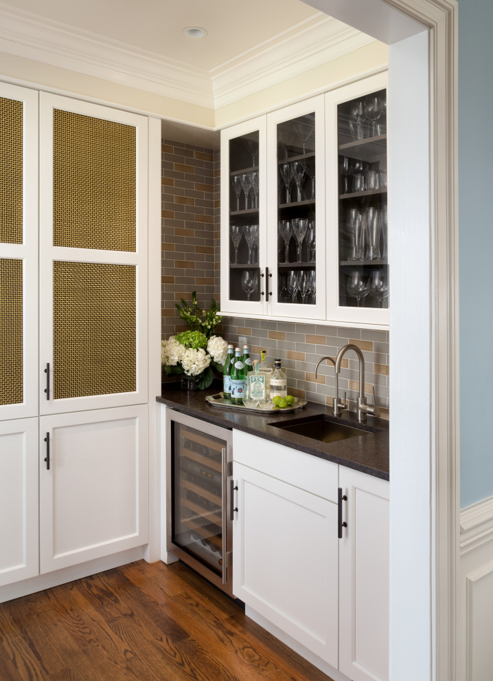 This is an example of a traditional single-wall wet bar in DC Metro with an undermount sink, glass-front cabinets, white cabinets, multi-coloured splashback, subway tile splashback, medium hardwood floors, brown floor and black benchtop.