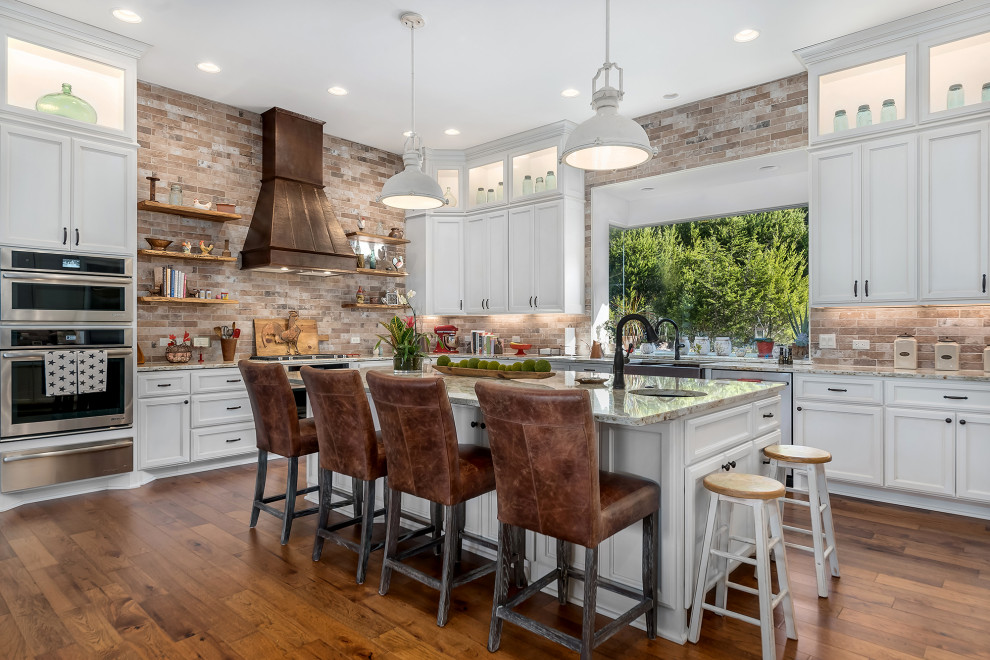Photo of a transitional u-shaped kitchen in Austin with a farmhouse sink, recessed-panel cabinets, white cabinets, granite benchtops, multi-coloured splashback, stone tile splashback, stainless steel appliances, medium hardwood floors, with island, brown floor and beige benchtop.
