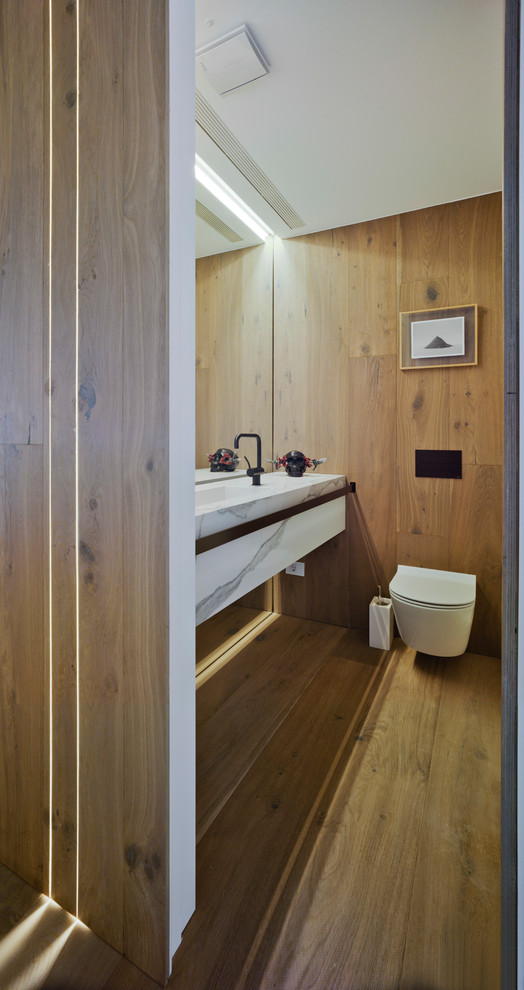Modelo de aseo contemporáneo pequeño con sanitario de pared, paredes blancas, suelo de madera en tonos medios, lavabo integrado, encimera de mármol y suelo marrón
