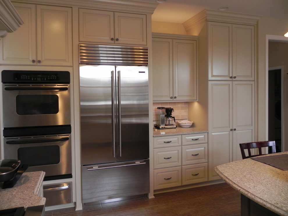 Example of a mid-sized transitional u-shaped light wood floor and brown floor eat-in kitchen design with an undermount sink, raised-panel cabinets, white cabinets, granite countertops, beige backsplash, ceramic backsplash, stainless steel appliances and an island