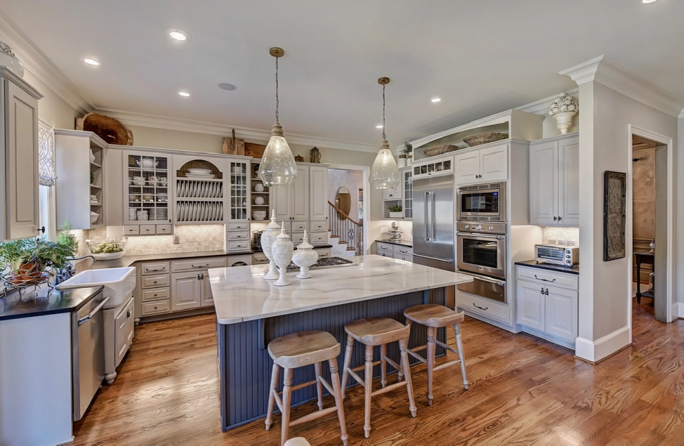 Large eat-in kitchen in Charlotte with a farmhouse sink, raised-panel cabinets, beige cabinets, quartzite benchtops, grey splashback, ceramic splashback, stainless steel appliances, medium hardwood floors, with island, brown floor and white benchtop.