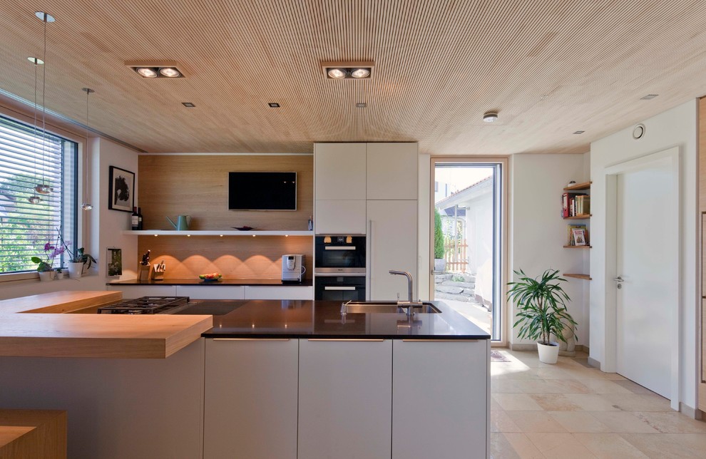 Inspiration for a mid-sized contemporary galley open plan kitchen in Munich with an integrated sink, flat-panel cabinets, white cabinets, brown splashback, timber splashback, black appliances, a peninsula, beige floor and black benchtop.