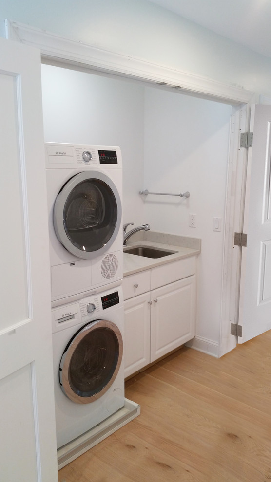 This is an example of a mid-sized country single-wall laundry cupboard in New York with an undermount sink, raised-panel cabinets, white cabinets, laminate benchtops, white walls, light hardwood floors, a stacked washer and dryer and beige floor.