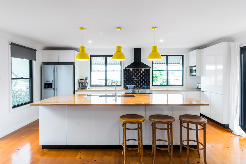 This is an example of a contemporary l-shaped kitchen in Brisbane with a drop-in sink, flat-panel cabinets, white cabinets, wood benchtops, black splashback, subway tile splashback, stainless steel appliances, medium hardwood floors, with island, brown floor and brown benchtop.
