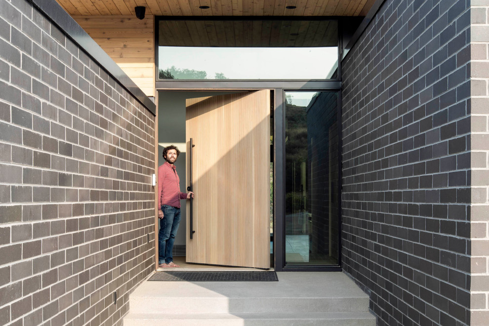 Exemple d'une porte d'entrée moderne avec sol en béton ciré, une porte pivot, une porte en bois clair, un sol gris, un plafond en bois et un mur en parement de brique.