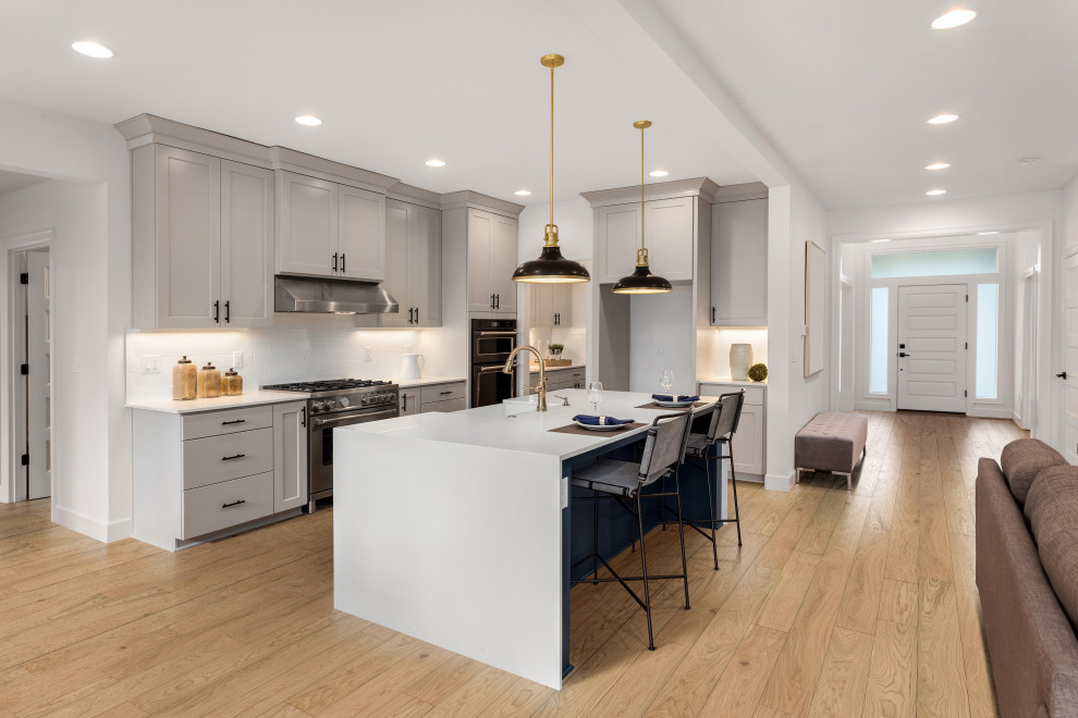 Photo of a contemporary l-shaped open plan kitchen in Los Angeles with a drop-in sink, flat-panel cabinets, grey cabinets, granite benchtops, white splashback, subway tile splashback, stainless steel appliances, light hardwood floors, with island, brown floor and white benchtop.