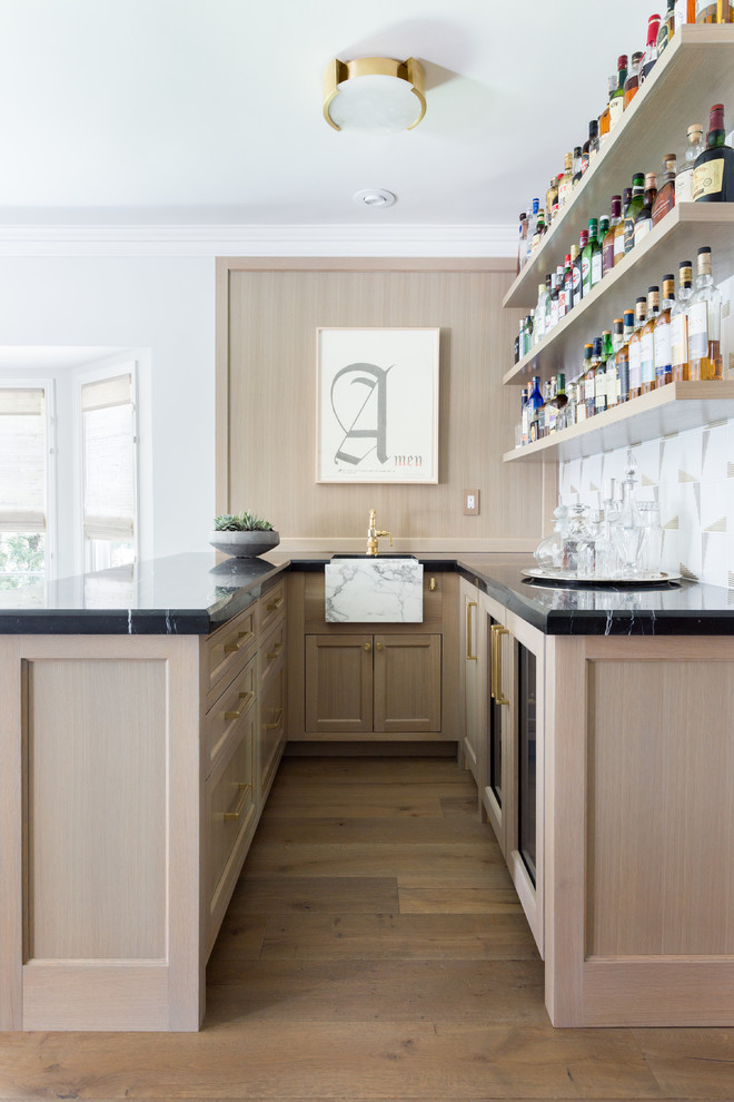 Inspiration for a transitional u-shaped seated home bar in Los Angeles with an undermount sink, shaker cabinets, white splashback, light hardwood floors and black benchtop.