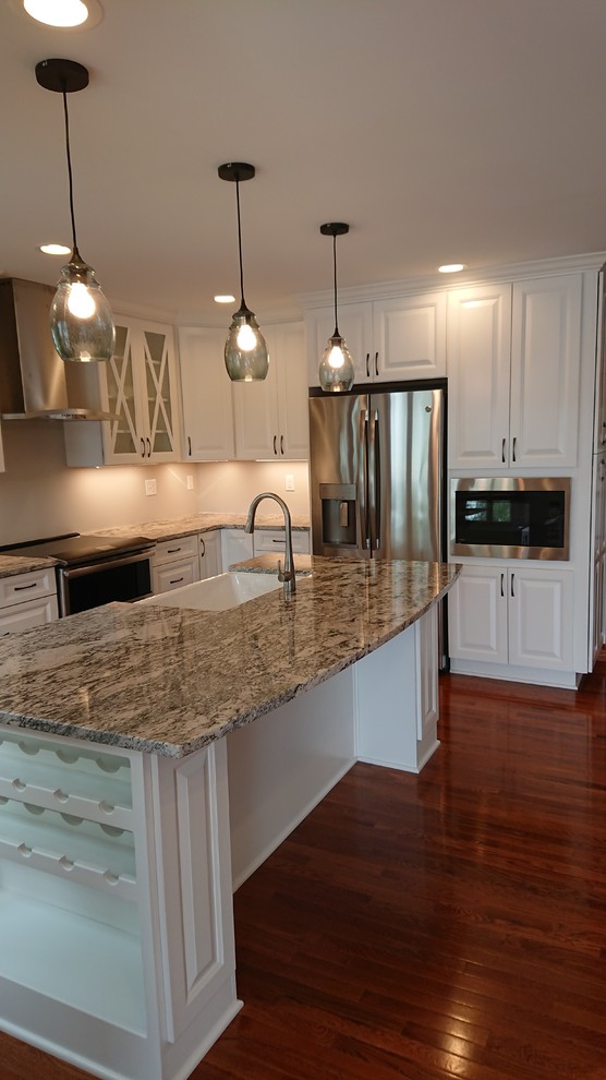 Photo of a mid-sized beach style l-shaped open plan kitchen in Baltimore with a farmhouse sink, raised-panel cabinets, white cabinets, granite benchtops, stainless steel appliances, medium hardwood floors, with island, brown floor and multi-coloured benchtop.