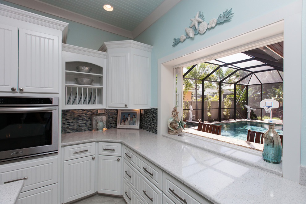 Photo of a beach style u-shaped eat-in kitchen in Tampa with a double-bowl sink, beaded inset cabinets, white cabinets, quartz benchtops, multi-coloured splashback, glass sheet splashback, stainless steel appliances, porcelain floors and with island.
