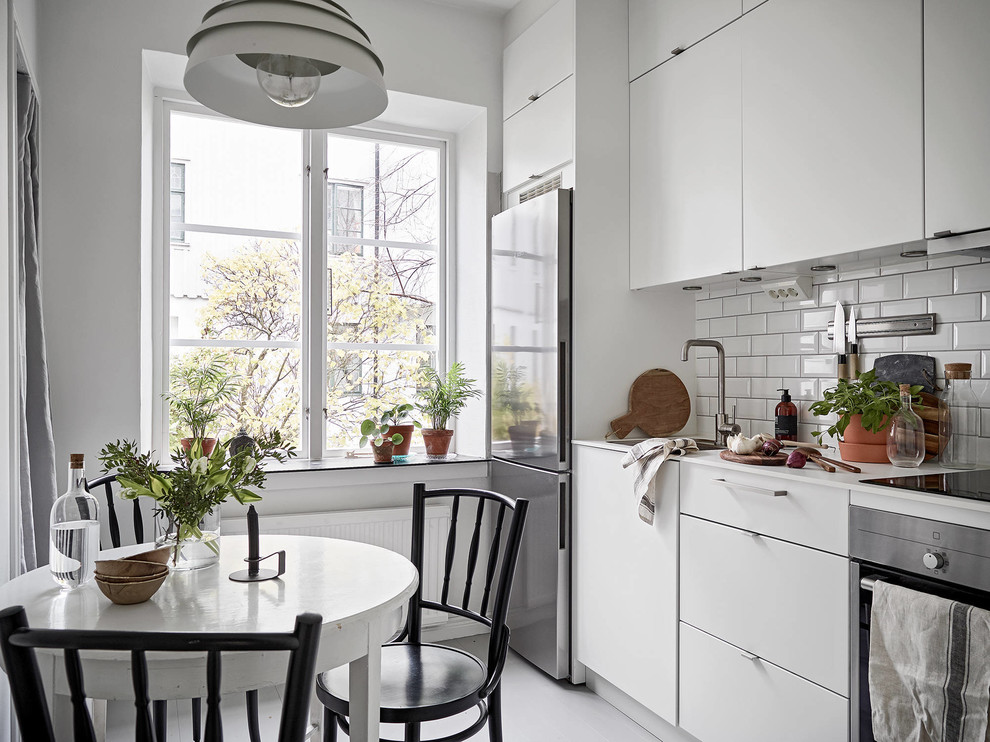This is an example of a scandinavian single-wall eat-in kitchen in Gothenburg with a single-bowl sink, flat-panel cabinets, white cabinets, white splashback, subway tile splashback, stainless steel appliances, white floor and white benchtop.