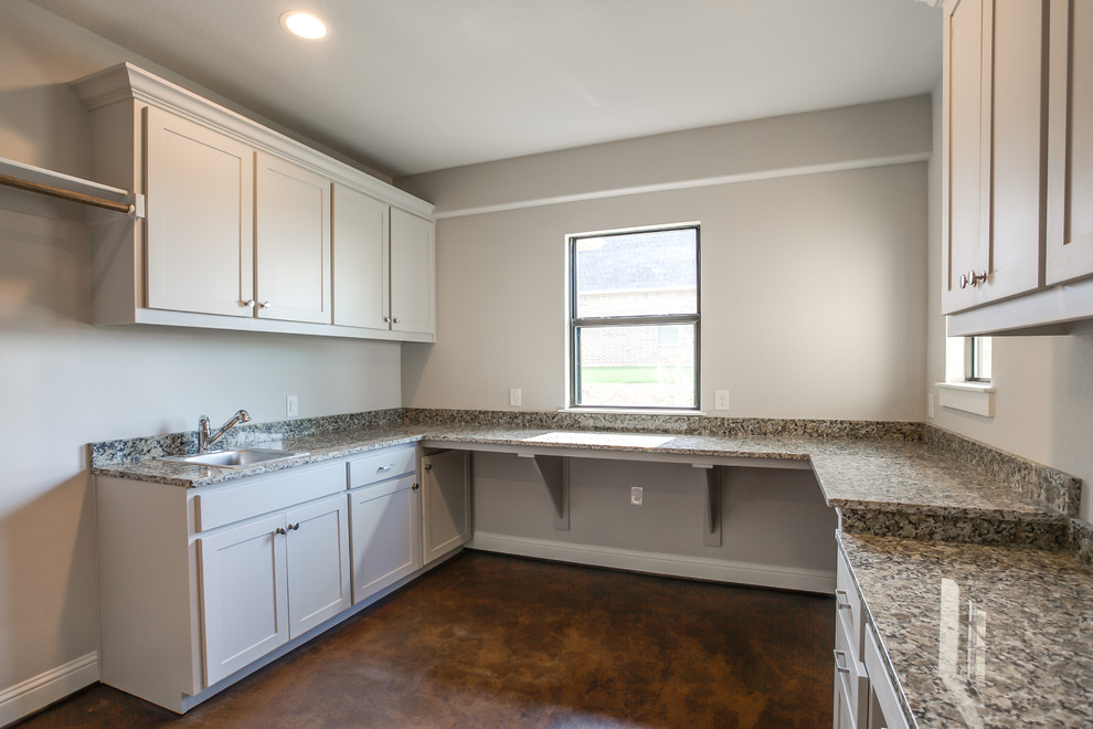 Photo of a mid-sized country u-shaped utility room in Dallas with a drop-in sink, shaker cabinets, grey cabinets, granite benchtops, grey walls, concrete floors, a stacked washer and dryer and brown floor.