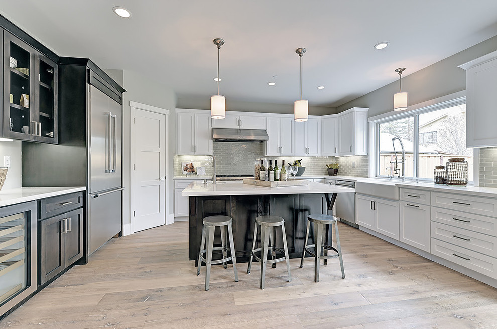 Large transitional u-shaped eat-in kitchen in San Francisco with a farmhouse sink, shaker cabinets, white cabinets, grey splashback, glass tile splashback, stainless steel appliances, light hardwood floors, with island and solid surface benchtops.