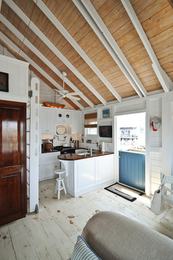 This is an example of a small beach style u-shaped separate kitchen in Boston with a farmhouse sink, beaded inset cabinets, white cabinets, wood benchtops, white splashback, black appliances, light hardwood floors and a peninsula.