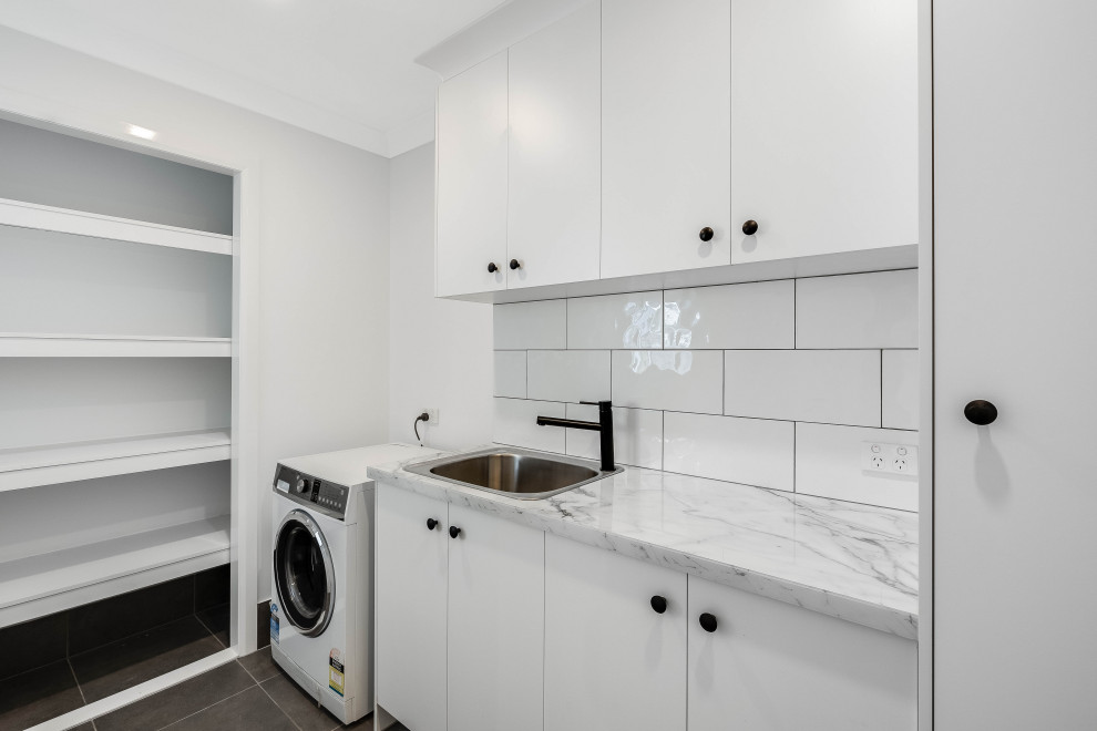 Mid-sized transitional dedicated laundry room in Other with a drop-in sink, ceramic splashback, white walls and grey floor.